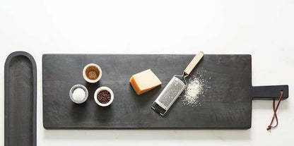 top view of the black charcuterie board with cheese and slicer and bowls of spices displayed on it next to a black oval tray on a white background