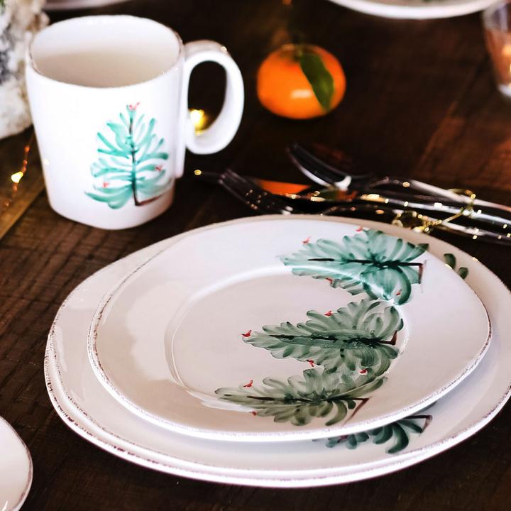 table setting on wooden tabletop with a dinner plate, salad plate, and mug that have trees painted on them. Silverware and an orange in the background.