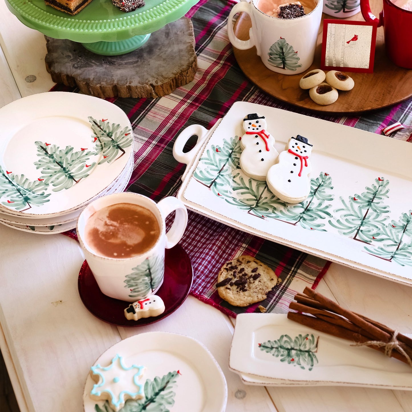 holiday platters, plates, and mugs arranged on a wooden table with snowman cookies, hot cocoa, and cinnamon sticks.