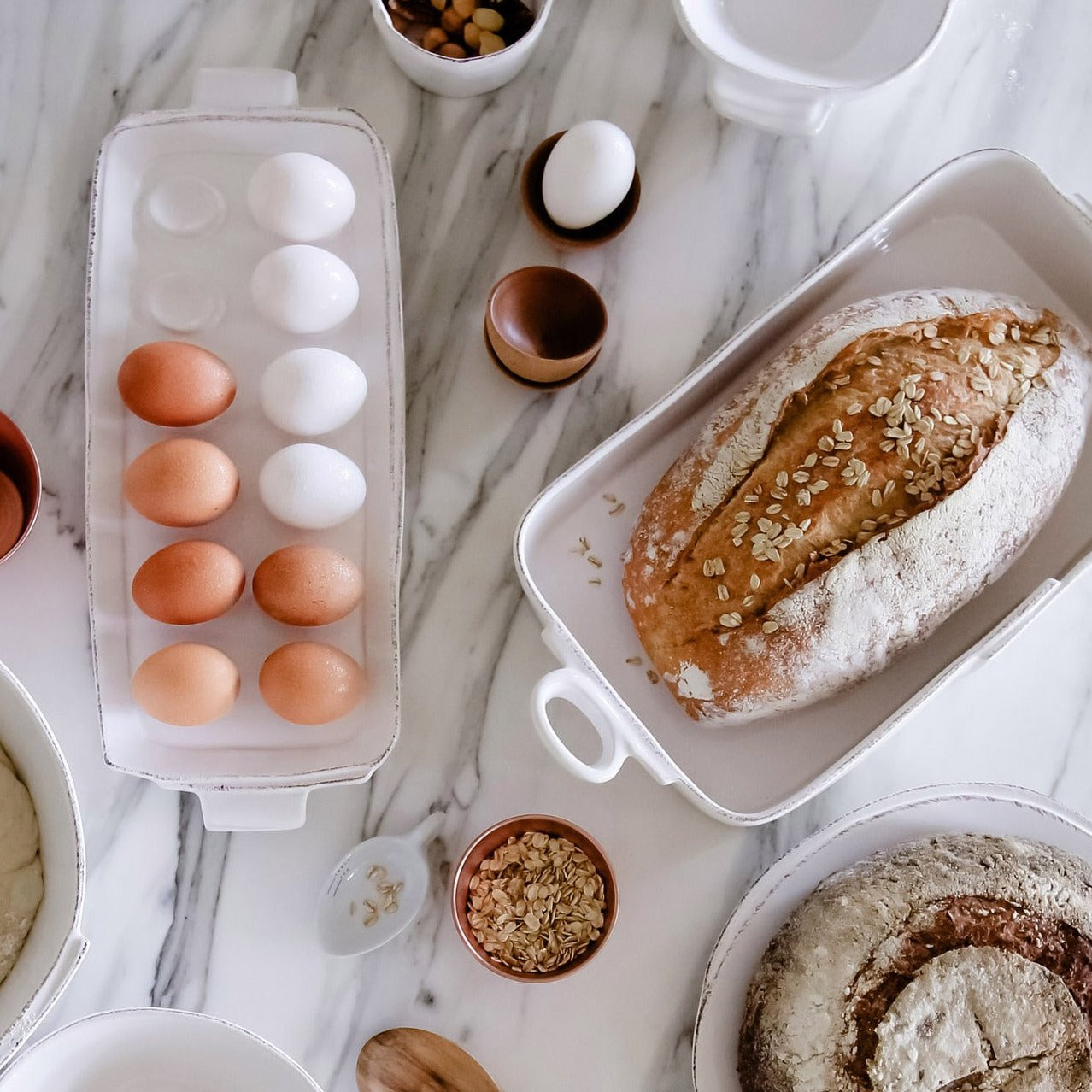 baking dish with loaf of bread, tray of eggs, and an array of small bowls arranged on a marble countertop.