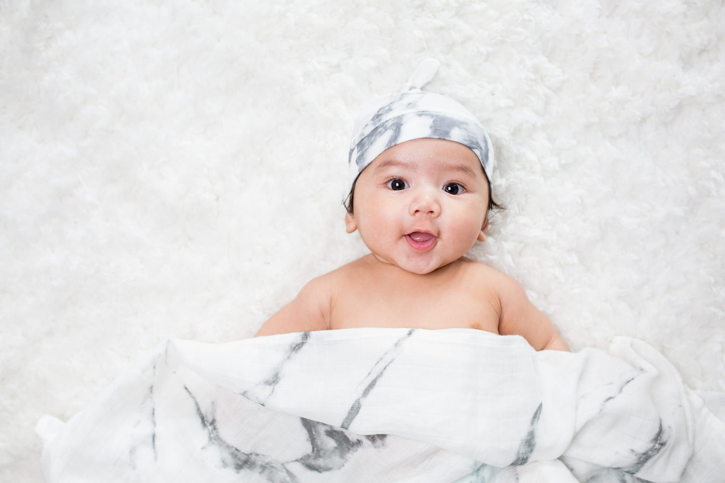 a baby wearing the marble hello world hat while laying under the marble swaddle on a white background