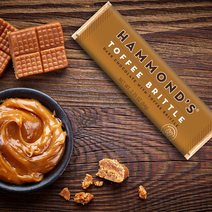 natural toffee brittle dark chocolate candy bar displayed in the package next to a bowl of toffee and pieces of brittle on a dark stained wood surface