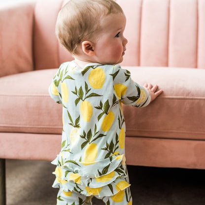 back view of a little girl wearing the lemon ruffle footed romper while holding on to a pink sofa