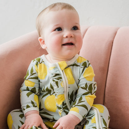 a little girl sitting on a pink sofa wearing the lemon ruffle footed romper