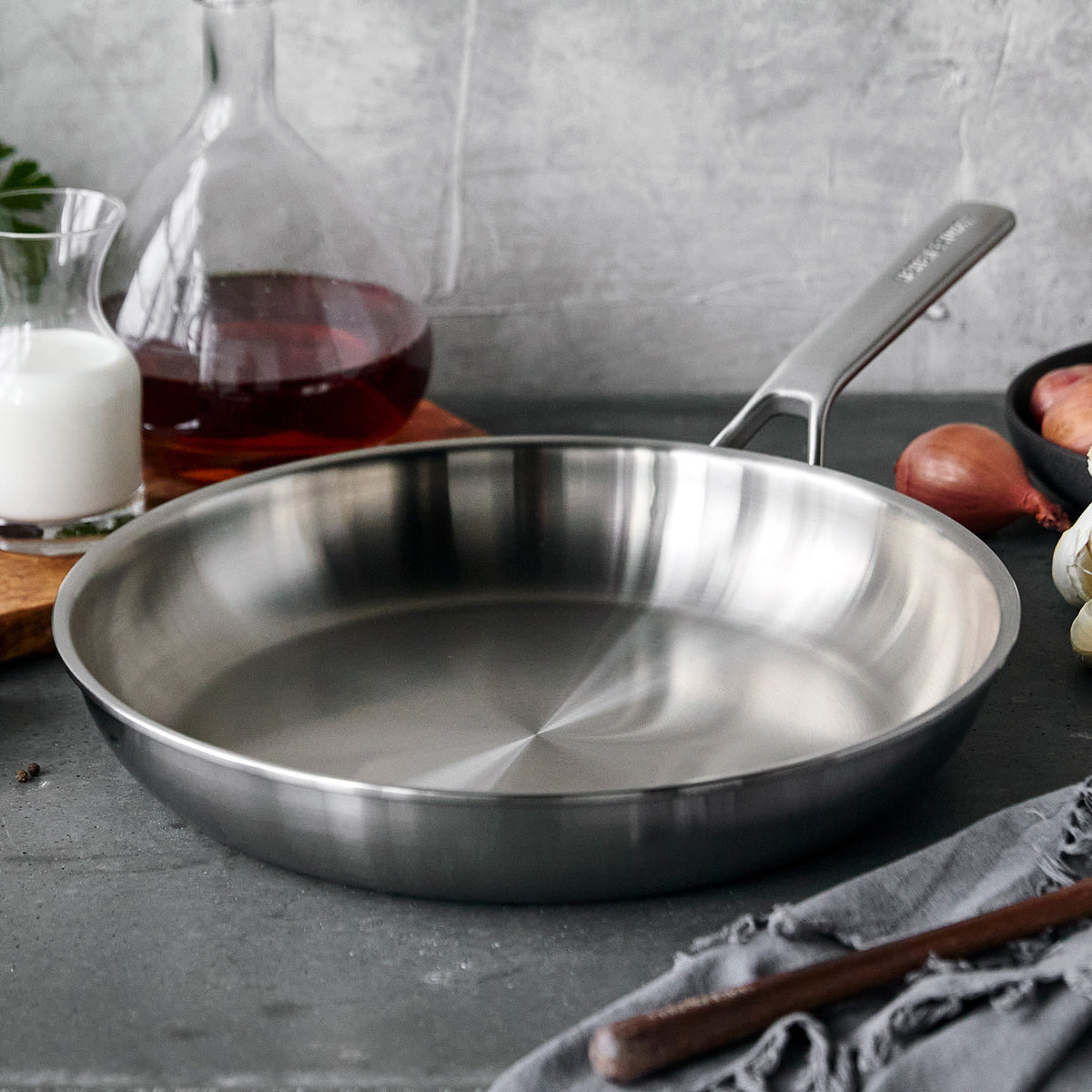 12 inch stainless steel frypan displayed on a dark gray surface next to a decanter, onions, and towel