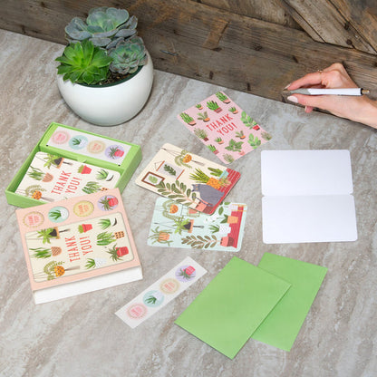open box of notecards, assorted cards with house plant graphics, and green envelopes on a marble countertop. hand holding pen prepares to write a note.