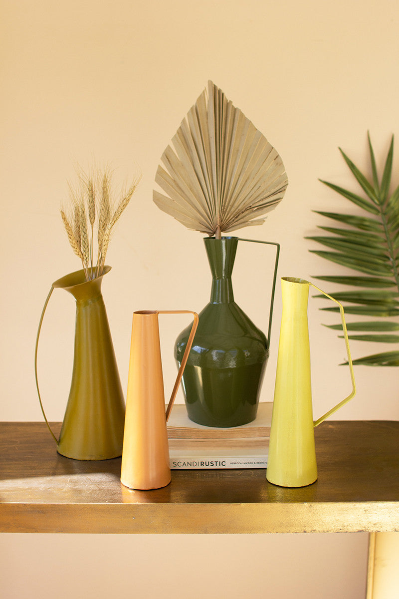 all four styles of metal vases with handles displayed on a wooden table displayed with dried palm leaves and wheat against a pale pink background