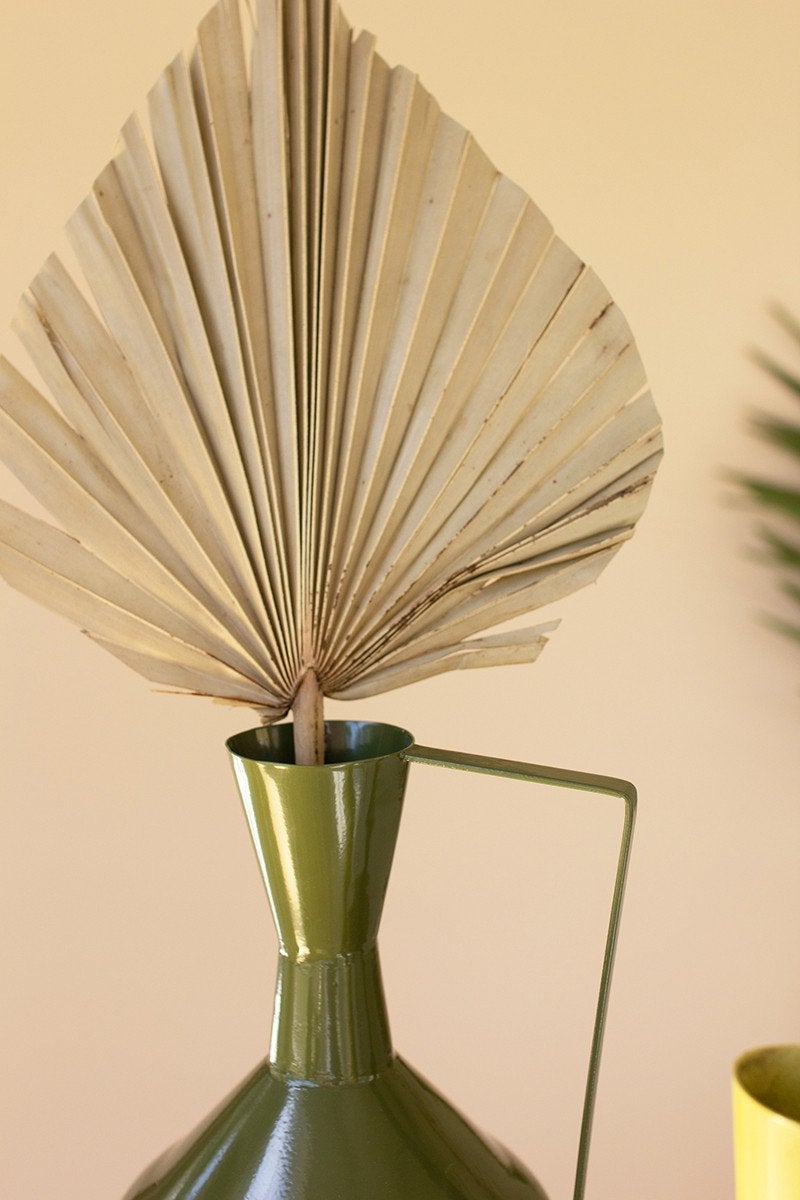close up view of the green metal vase with handle filled with a dried palm leaf against a pale pink background