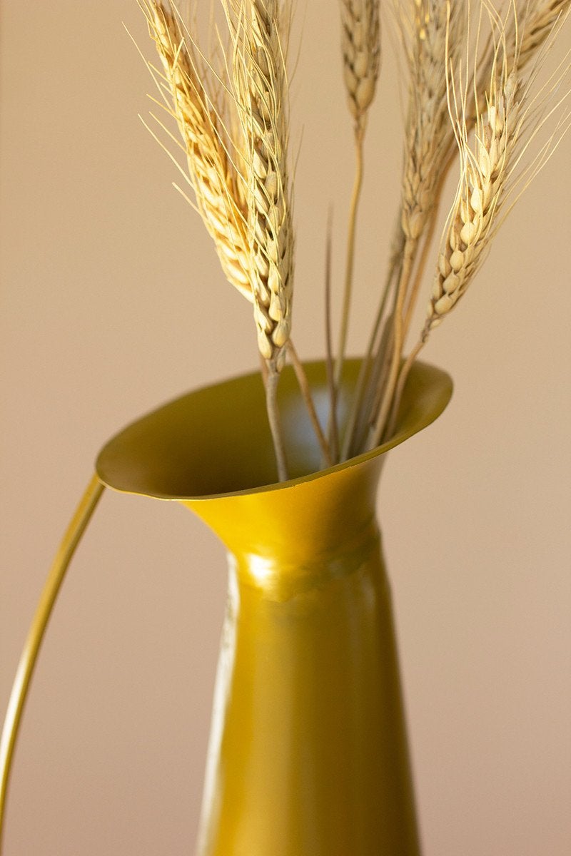 close up view of the mustard metal vase with handle filled with dried wheat against a pale pink background
