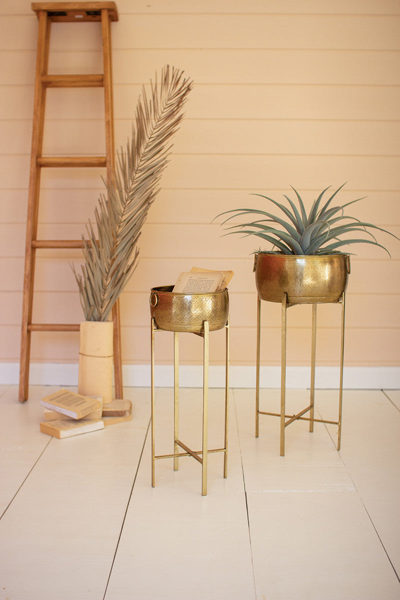 2 sizes of brass finished pots on stands, larger has plant in it, smaller has papers in it.