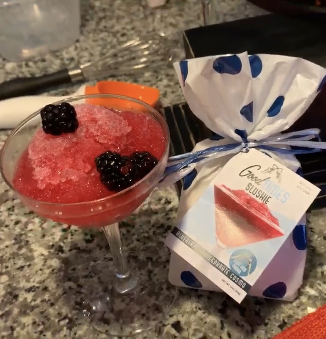 blueberry pomegranate cosmo slushie displayed in a glass with blackberries beside the container bag on a granite countertop