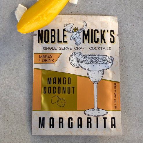noble mick's single serve packet of Mango Coconut margarita mix on a bar top with a mango wedge and coconut shavings.