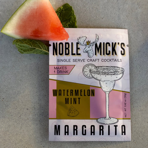 noble mick's single serve packet of Watermelon Mint margarita mix on a bar top with a watermelon wedge and mint.
