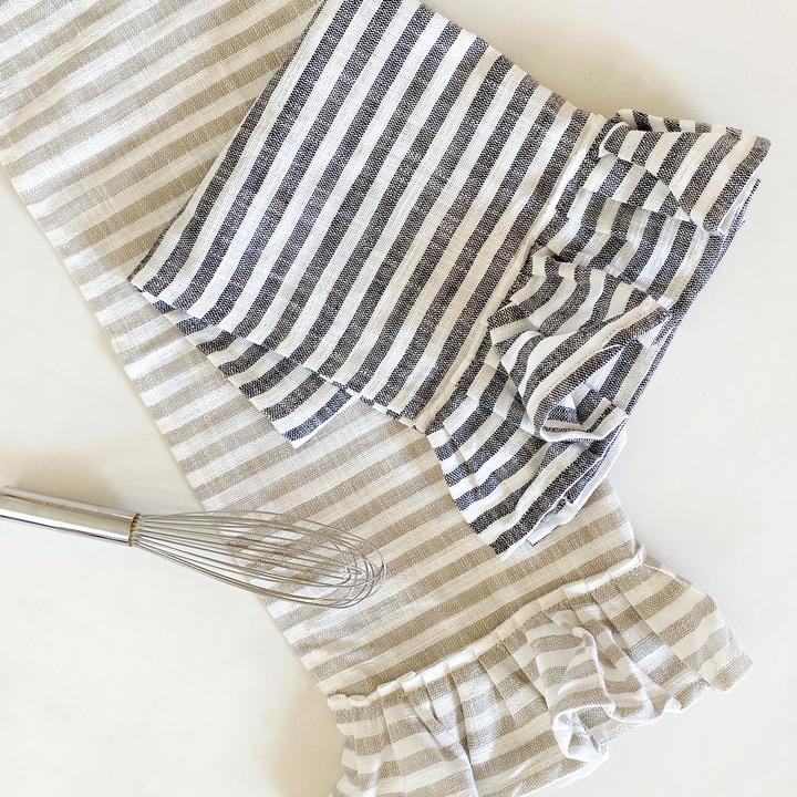 one natural and one black striped cotton tea towels with ruffles next to a whisk on a white background