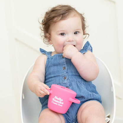 little girl sitting in a chair eating out of the hello snacks happy snacker on a mostly white background
