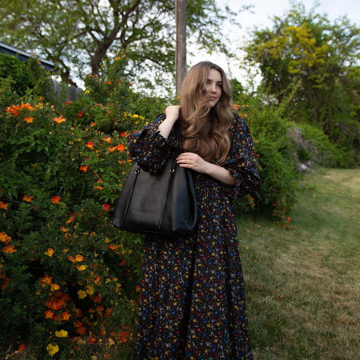 a woman standing in front of a row of flowering bushes outside with the black kingston tote bag over her shoulder
