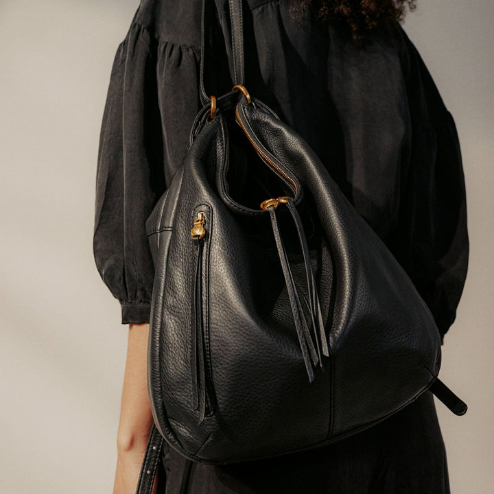 side view of a woman wearing the black merrin convertible backpack shoulder bag against a light gray background