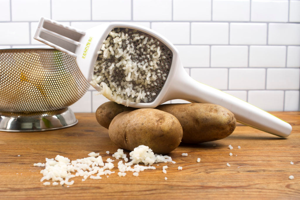  potato ricer with riced potatoes coming out of it on wood counter with potatoes and colander.