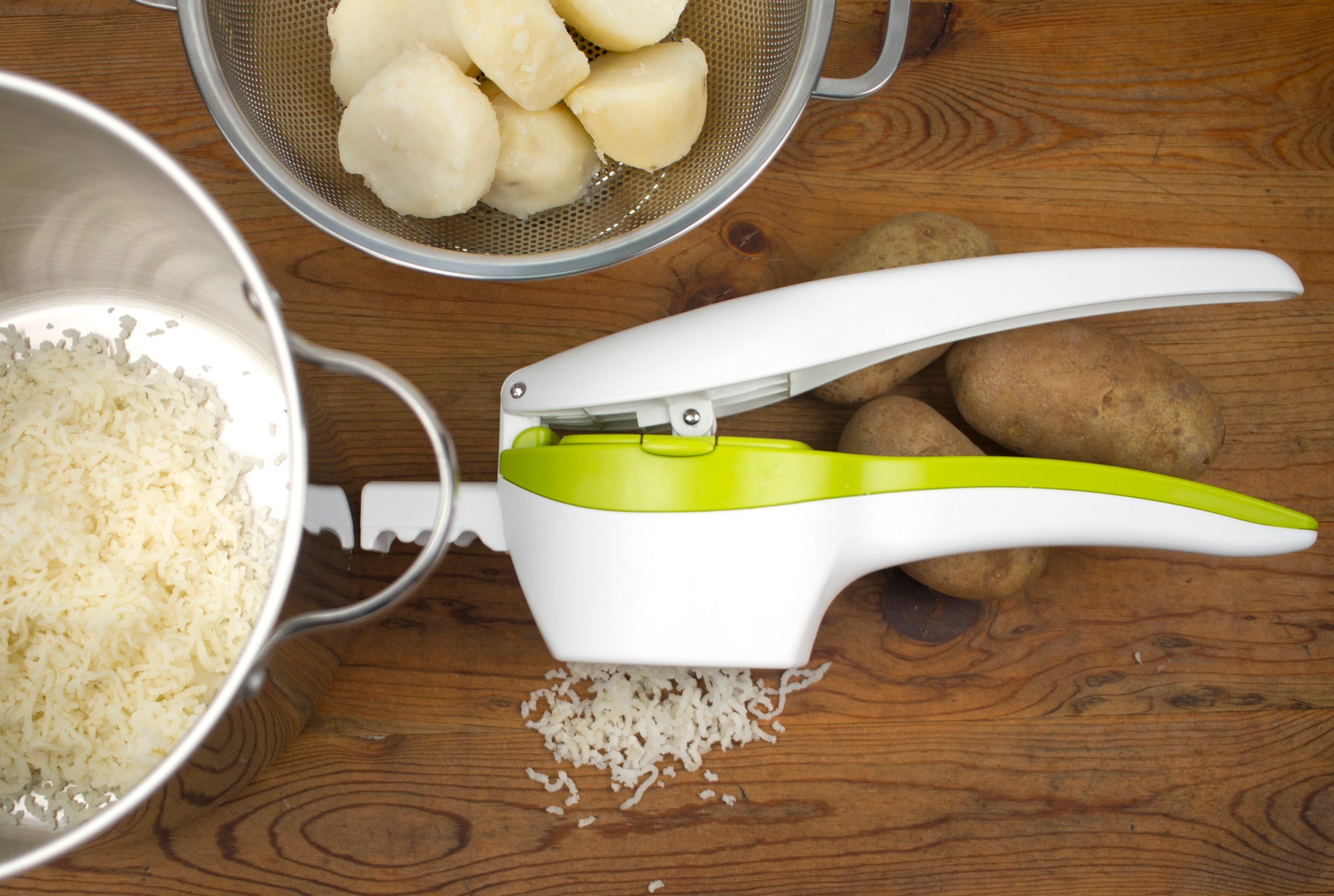 potato ricer on wood counter with potatoes, colander or peeled potatoes, and pan of riced potatoes.