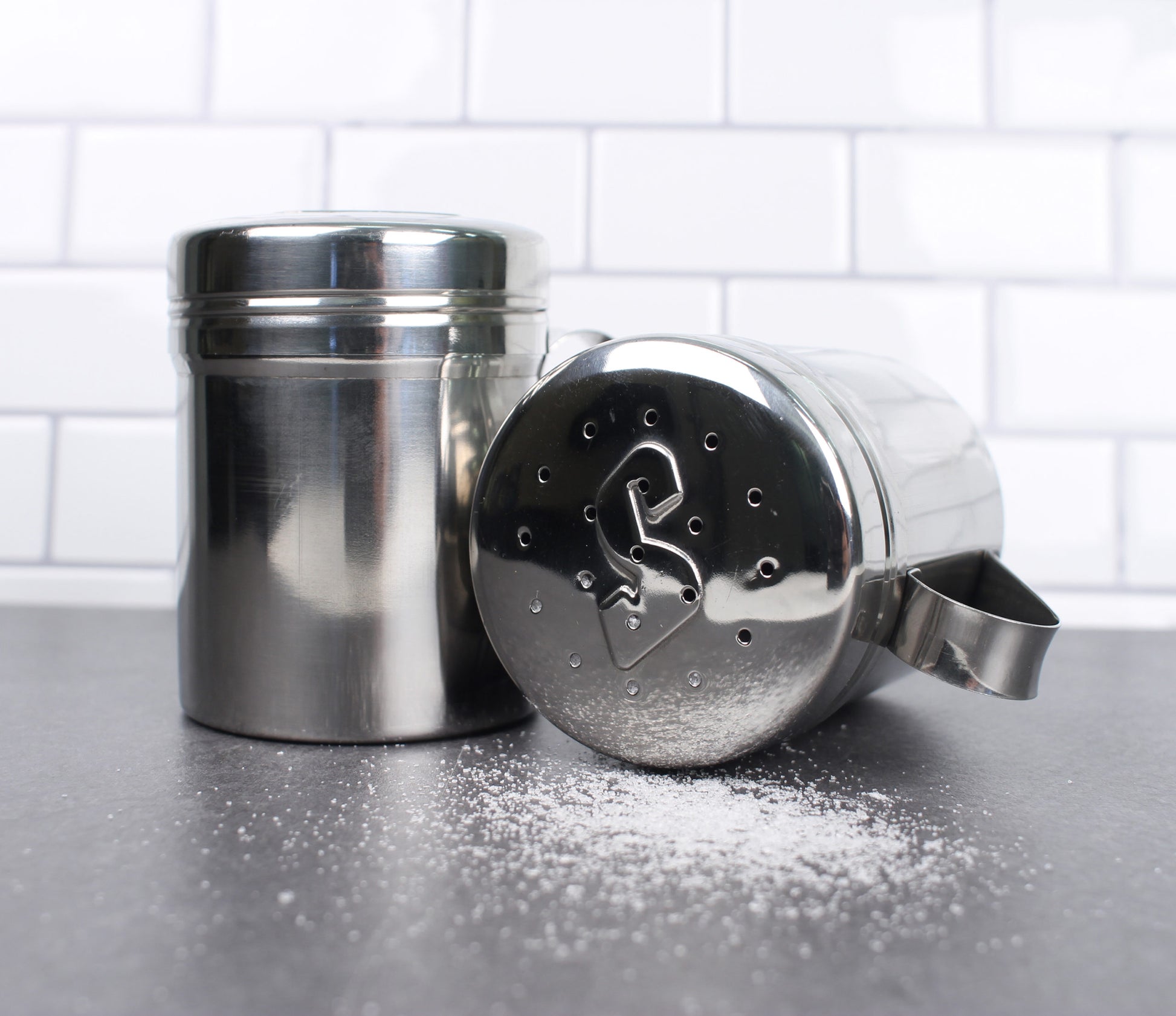 shakers on grey counter top with salt shaker is laying down with salt shaken out.