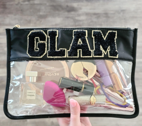 hand holding clear nylon bag with black trim, chenille patches that spell "glam", and filled with make-up.