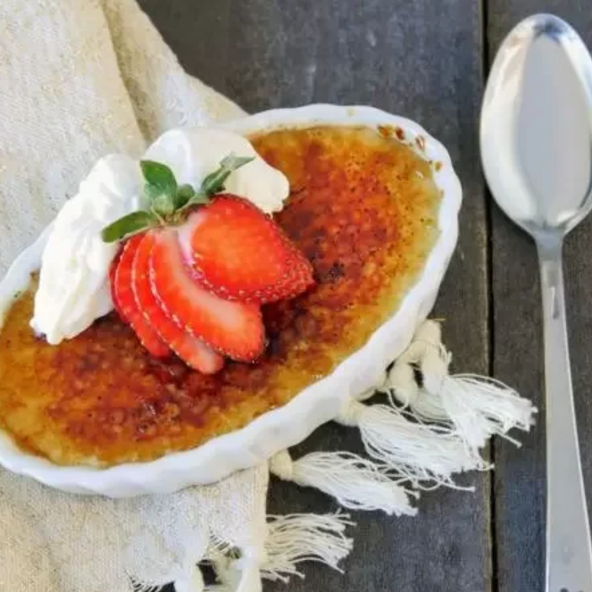 oval dish with creme brulee topped with strawberries and cream on wooden tabletop with fringed cloth and spoon.