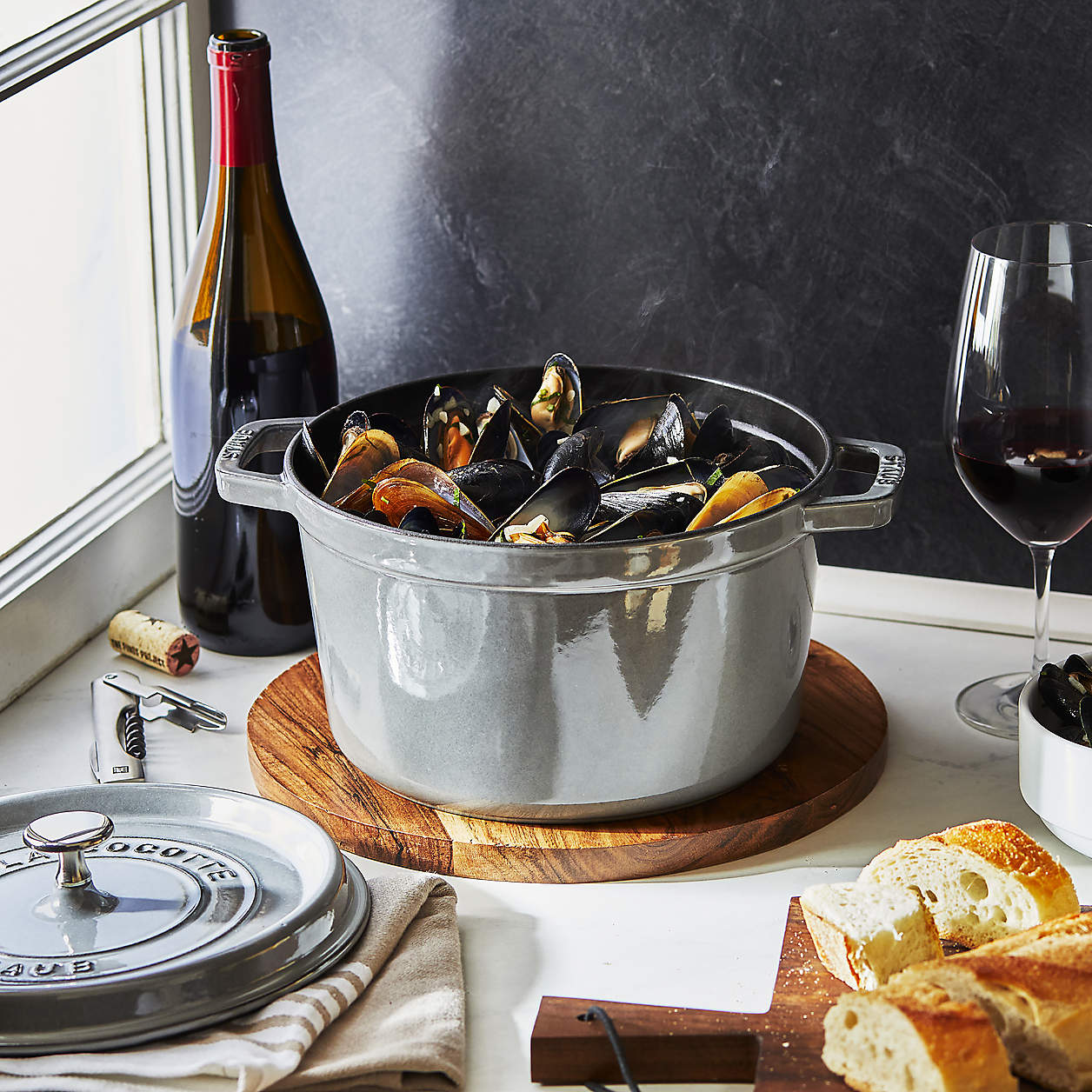 dutch oven on tabletop filled with oysters. glass and bottle of wine and bread on cutting board also on the table. A window lights the table set in front of a black back ground.
