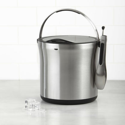 stainless steel ice bucket with tongs attached to the side and ice cubes next to it.