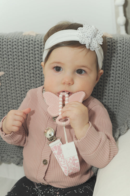 a baby girl chewing on the butterfly brush toothbrush teether against a gray blanket while sitting in a white chair