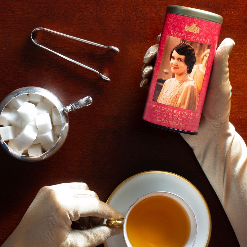 a woman wearing white gloves while holding a canister of lady cora's evening herbal tea next to a cup of tea, tea tongs and bowl of sugar cubes