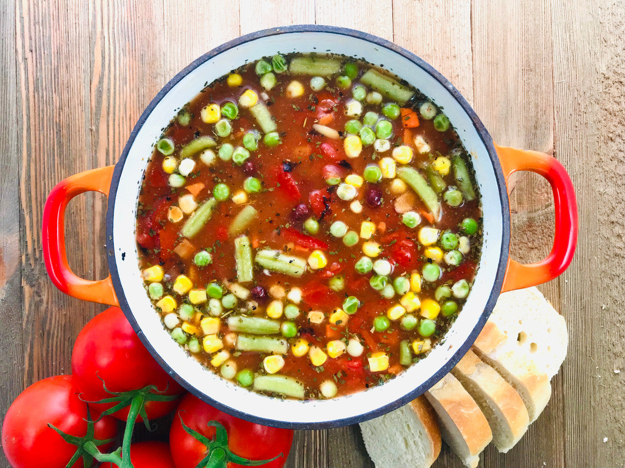 vegetable orzo soup displayed in a pot next to tomatoes and sliced bread on a wooden surface