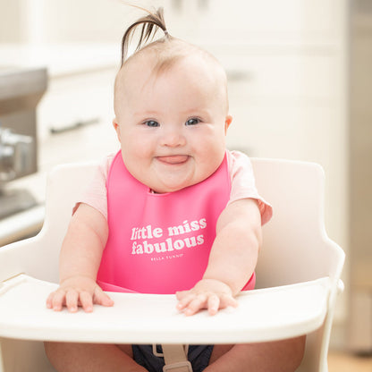 a little girl sitting in a highchair smiling with her tung out and wearing the little miss fabulous wonder bib
