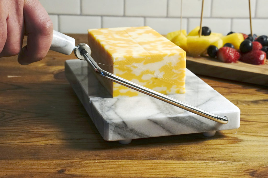 cheese slicer on wood counter with block of cheese being sliced, sliced fruit on background.