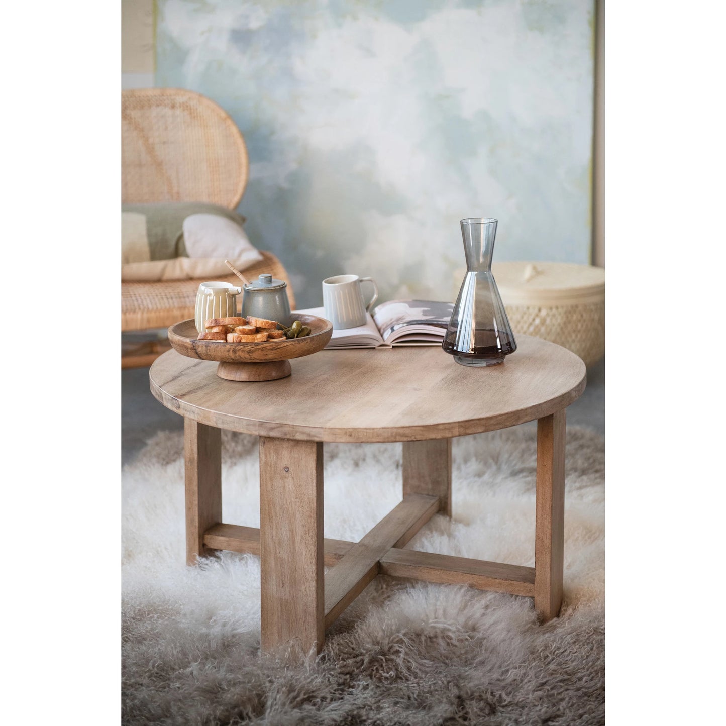 decanter set on a round coffee table with an open book, wooden bowl and mugs, table is set on a faux fur rug.