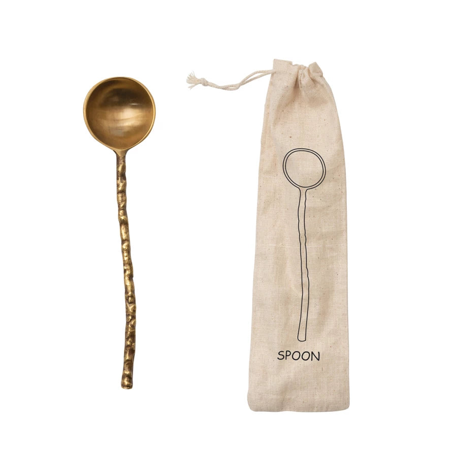 brass spoon with hammered and slightly curved handle next to a muslin bag on a white background.