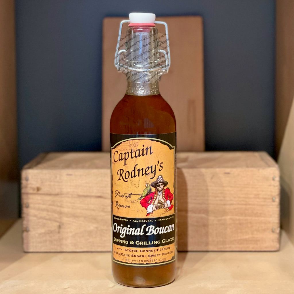 bottle of Captain Rodney's Original Boucan Glaze on a table with wooden boxes in the background.
