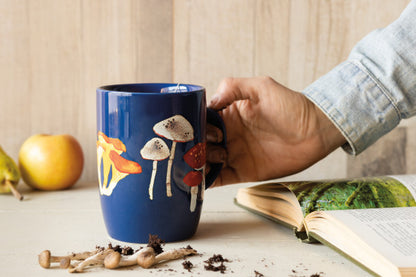 a persons hand picking up the field mushrooms tall mug off of a white table next to a book, yellow apples, and mushrooms