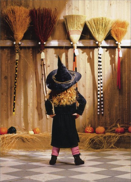 front of card has a little girl in a witch costume facing a wall of brooms in a barn