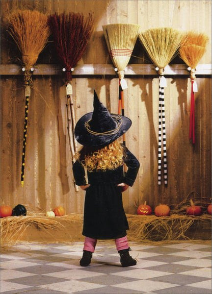 front of card has a little girl in a witch costume facing a wall of brooms in a barn