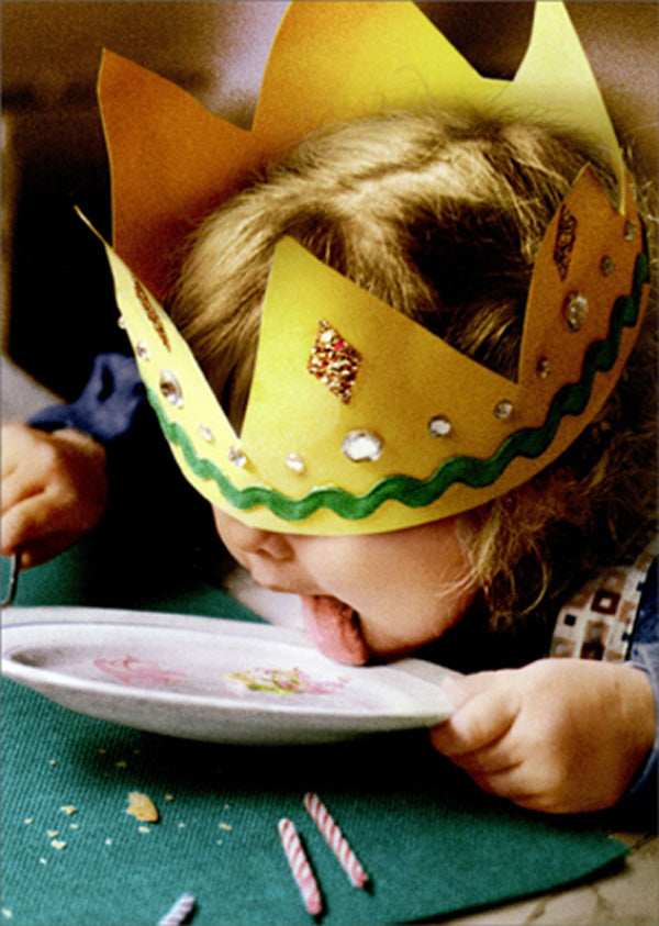 front of card is a photograph of a small childing licking her plate with a crown on her head and birthday candles on the table