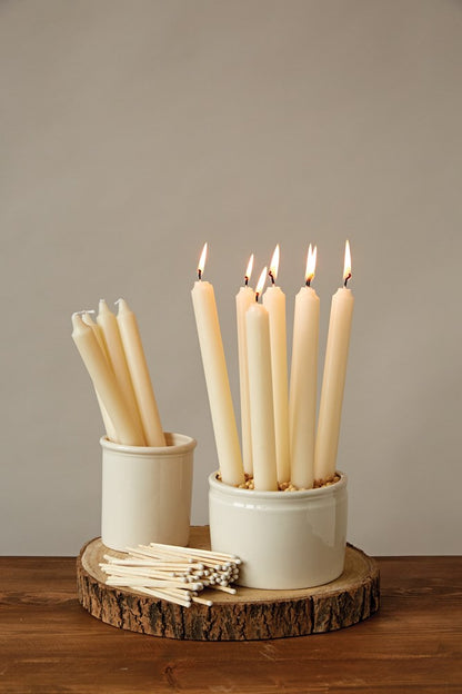 a display of ivory tall tapers burning in a bowl and several in a cup on a round wood cookie slab on a dark stained table against a gray background
