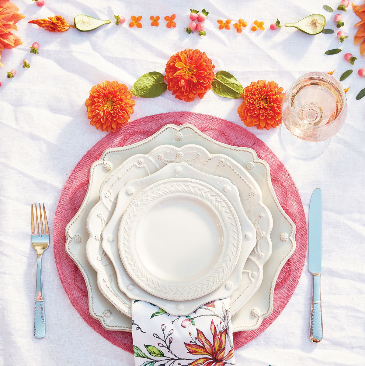 four piece place setting of juliska dinnerware displayed on a pink charger with flatware, floral napkin and orange flowers on a white tablecloth