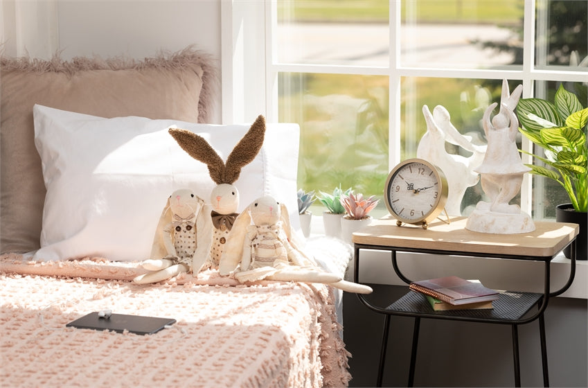 bedroom scene with desk clock on a side table and stuffed animal bunnies on the bed leaning against the pillows.