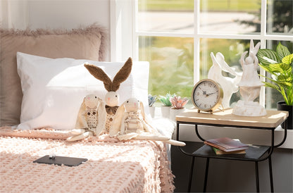 bedroom scene with desk clock on a side table and stuffed animal bunnies on the bed leaning against the pillows.