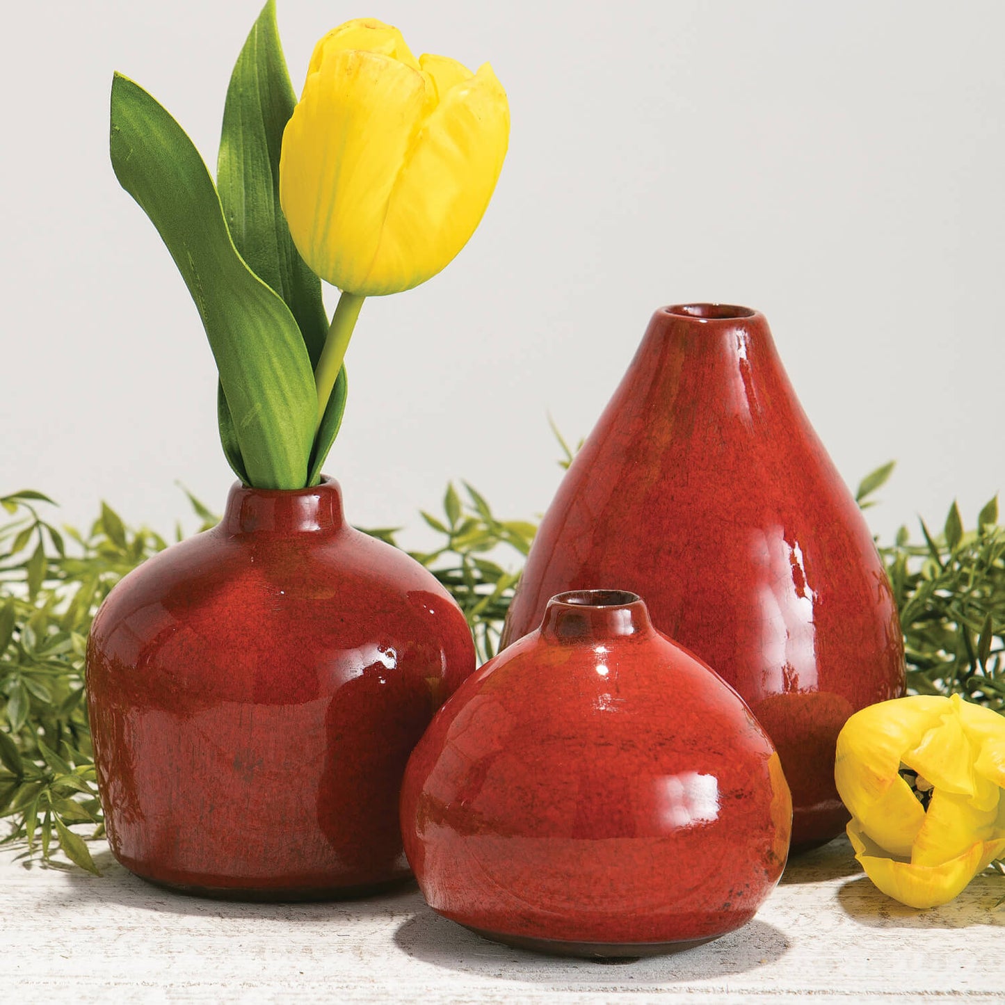 all three sizes of dappled red vases with a yellow tulip in the medium one and a yellow tulip laying next to them 