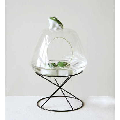 glass dome on top of wire stand with 2 ceramic frogs inside and one on top.