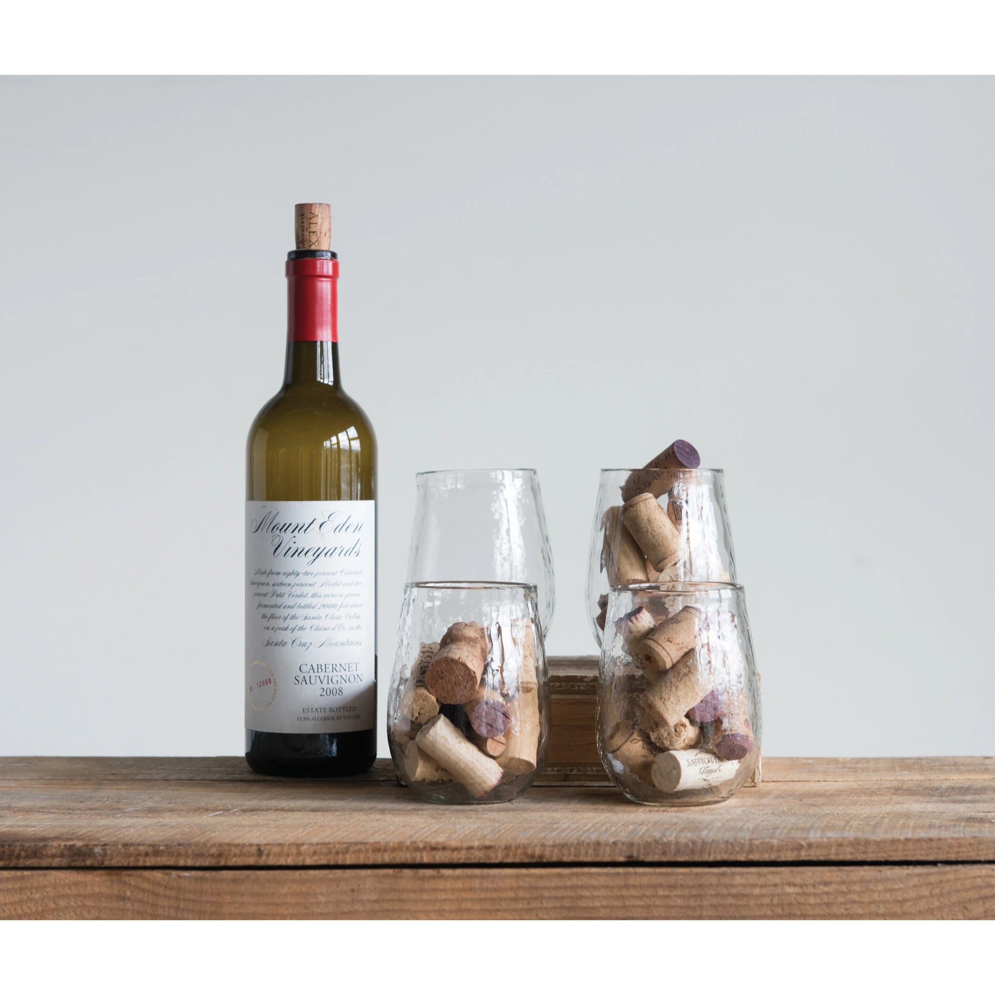 four recycled glass stemless wine glasses displayed with corks inside next to a wine bottle on a rustic wooden surface against a light blue background