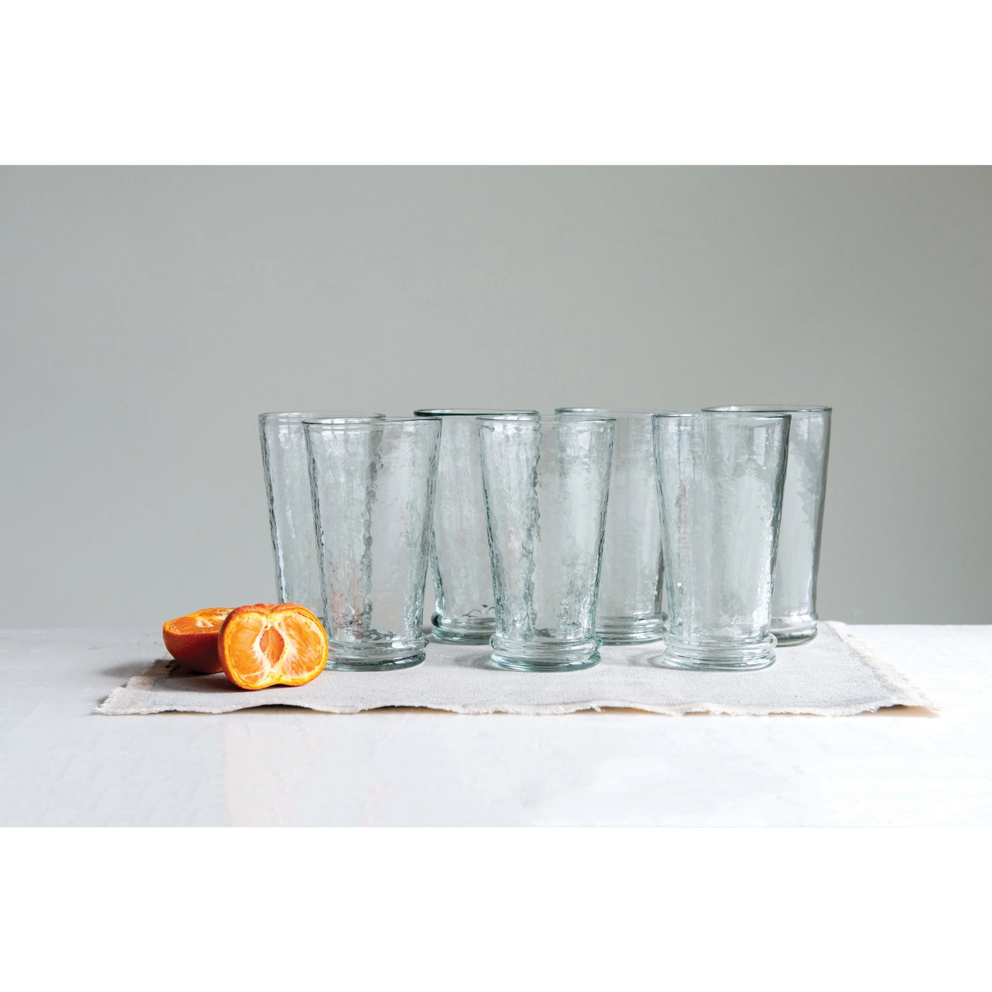 multiple recycled glass drinking glasses displayed next to sliced fruit on a placemat against a light gray wall
