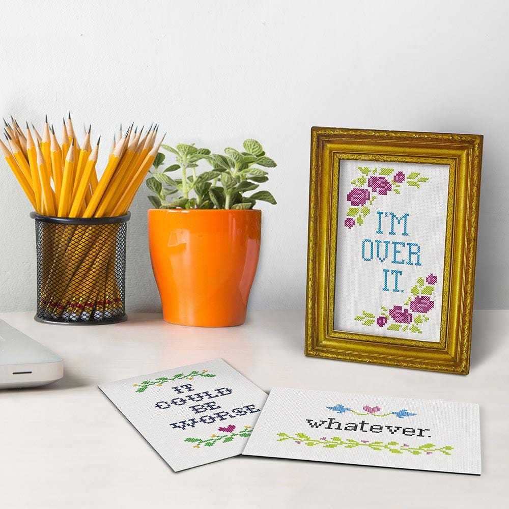 gold frame with text "i'm over it" on a desk with 2 other cards with text "it could be worse" and "whatever" set nest to a small plant and a jar of pencils.
