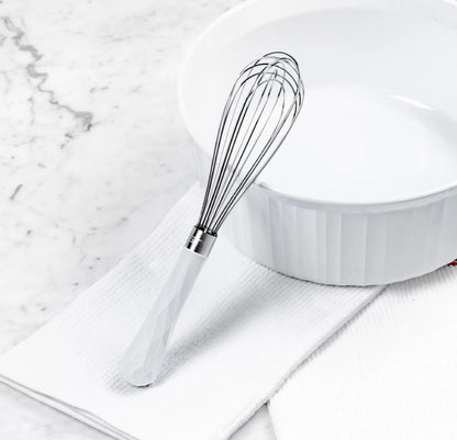 mini whisk laying on a white dishtowel leaning on a mixing bowl on a white countertop.